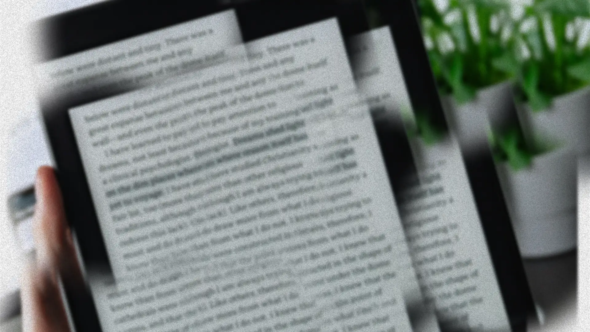 A blurred close-up of a hand holding a Kindle e-reader, with green foliage visible in the background.